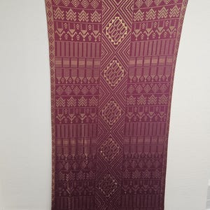 May include: A long, rectangular textile with a deep burgundy base and intricate gold geometric patterns. The design includes repeating shapes and lines, creating a symmetrical and ornate appearance. The fabric is hanging vertically.