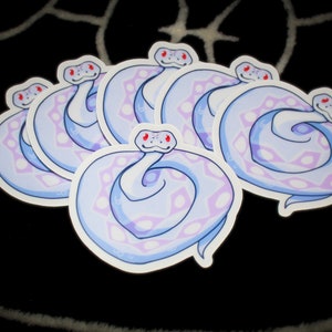 Op de afbeelding: Een verzameling stickers met een gestileerd slangenontwerp. De slangen zijn voornamelijk lichtblauw met lavendel- en witte patronen, rode ogen en een witte omtrek. De stickers zijn gerangschikt op een zwart oppervlak.