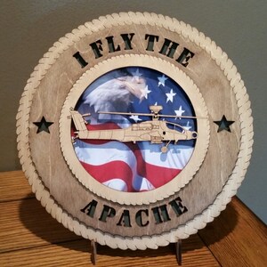 May include: A wooden plaque with a rope border and the text "I Fly The Apache" in green letters. The plaque has a circular cutout with an American flag and a helicopter inside.