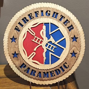 May include: A wooden round sign with a rope border. The sign features a red and blue firefighter and paramedic symbol with a ladder and a snake. The words "Firefighter Paramedic" are written around the symbol in a rope-like font. There are four stars around the symbol.