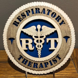 May include: A wooden sign with the words "RESPIRATORY THERAPIST" and "R T" in a circular design. The sign features a blue background with a medical symbol and a rope-like border. The sign is made of layered wood.