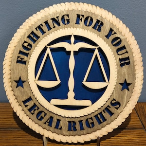 May include: A wooden round sign with a rope border. The sign has a blue background with a white scale of justice and the text "FIGHTING FOR YOUR LEGAL RIGHTS" in a brown font.