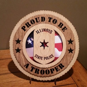 May include: Wooden plaque with a rope border, featuring the text "Proud to be a Trooper" and a cutout of the Illinois State Police badge with a star and the state outline. The badge is superimposed on a cutout of the American flag.