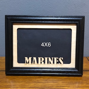 May include: Black wooden picture frame with a light wood interior and a black backing. The frame is labeled "4X6" and has the word "MARINES" cut out at the bottom. The frame is sitting on a wooden surface.