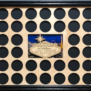 May include: A wooden coin display frame with 25 circular slots for coins. The frame has a black border and a cutout in the center that displays a Las Vegas welcome sign with a cityscape background.