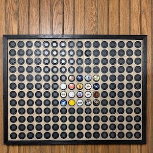 May include: A framed display board with a grid of black bottle cap holders. A selection of colorful bottle caps are displayed in the center, surrounded by the black holders. The frame is black, and the background is a light wood grain.