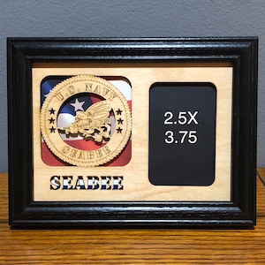 May include: A wooden picture frame with a black border. It features a cutout for a 2.5 x 3.75 inch photo and a circular emblem with the text "U.S. NAVY SEABEE." The emblem is set against a backdrop of the American flag.