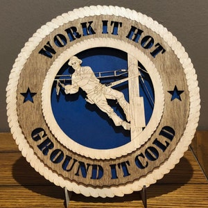May include: A wooden sign with a rope border and the text "Work It Hot Ground It Cold". The sign features a silhouette of a lineman working on a power pole.