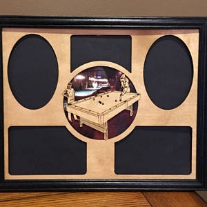 May include: A black wood picture frame with a light wood background. The frame has six openings for pictures. The center opening shows a pool table with two people playing pool. The other openings are empty.