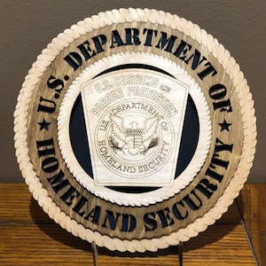 May include: A wooden plaque with a black and white image of the U.S. Department of Homeland Security seal. The seal features a bald eagle with a shield and the words "U.S. Customs and Border Protection" and "Department of Homeland Security". The plaque is surrounded by a rope-like border and the words "U.S. Department of Homeland Security" are written around the outside.