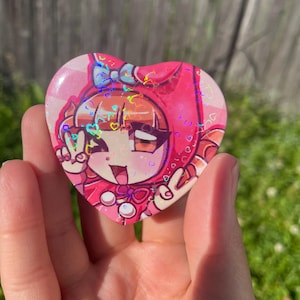 May include: A heart-shaped pin featuring a holographic finish with a repeating heart pattern. The design displays a cartoon character with brown hair, wearing a pink hood with a blue bow, making a peace sign with one hand against a pink background.