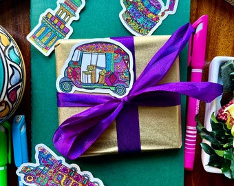 May include: A gold-wrapped gift tied with a purple ribbon, adorned with colorful stickers of a tuk-tuk, a teapot, and a tower. The scene includes a green notebook, pens, and decorative items, creating a vibrant, artistic display.