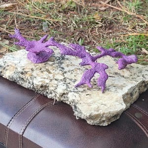 May include: A purple, organic-shaped metal sculpture sits on a piece of granite. The sculpture is abstract and resembles a branch or a tree.