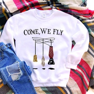 May include: White sweatshirt with the text "COME, WE FLY" printed in black. The graphic features three broomsticks with a red and white striped broom in the center.