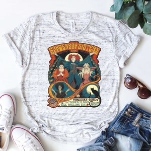 Pu&ograve; includere: Una maglietta bianca con una grafica in stile vintage delle sorelle Sanderson del film Hocus Pocus. La grafica presenta le tre streghe, un gatto nero e il testo "The Sanderson Sisters Back From The Dead Billy Butcherson Thackery Binx October 31, 1993 Salem All Hallows Eve".
