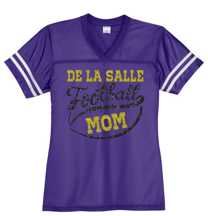 May include: Purple football jersey with white stripes on the sleeves. The jersey has gold glitter text that says "De La Salle Football Mom" with a black and gold glitter football graphic.