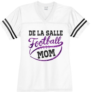 May include: White football jersey with black stripes on the sleeves. The jersey has a purple glitter design with the text "De La Salle Football Mom".