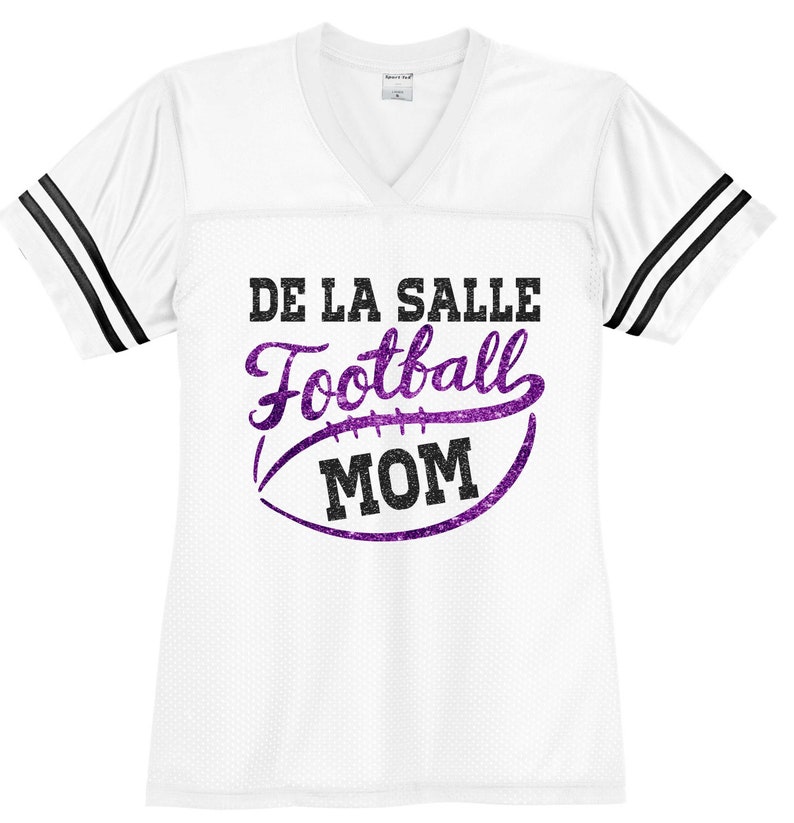 May include: White football jersey with black stripes on the sleeves. The jersey has a purple glitter design with the text "De La Salle Football Mom".