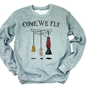 May include: A gray sweatshirt with the text "COME, WE FLY" printed on the front. The text is in black and is surrounded by three broomsticks, one of which is red and the other two are brown and beige.