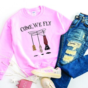May include: Pink sweatshirt with the text "Come, We Fly" and a graphic of three broomsticks. The sweatshirt is paired with blue jeans and white sneakers.