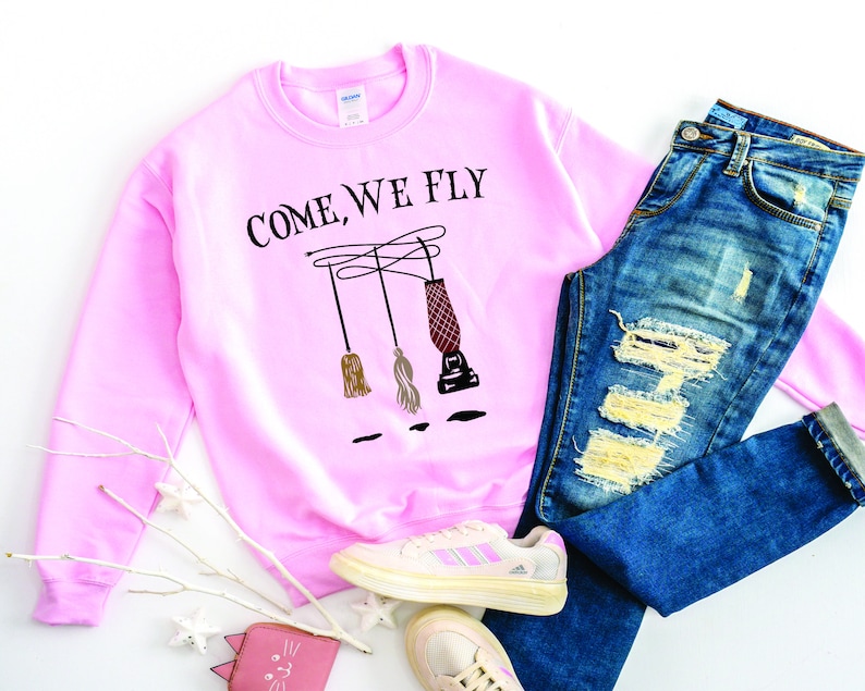 May include: Pink sweatshirt with the text "Come, We Fly" and a graphic of three broomsticks. The sweatshirt is paired with blue jeans and white sneakers.