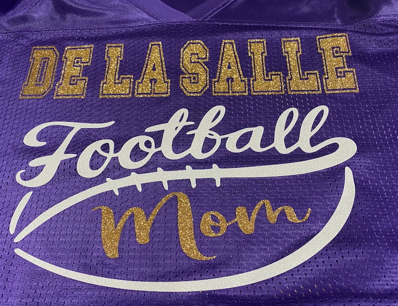 May include: Purple jersey with gold glitter lettering that reads "De La Salle Football Mom" with a white football graphic.
