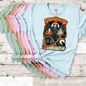 Pu&ograve; includere: Una pila di magliette di colore azzurro chiaro, rosa, pesca e verde con una stampa grafica delle sorelle Sanderson del film Hocus Pocus. La grafica include il testo "Sanderson Sisters" e "Hocus Pocus" con un gatto nero e un calderone di strega.