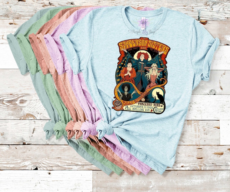 Pu&ograve; includere: Una pila di magliette di colore azzurro chiaro, rosa, pesca e verde con una stampa grafica delle sorelle Sanderson del film Hocus Pocus. La grafica include il testo "Sanderson Sisters" e "Hocus Pocus" con un gatto nero e un calderone di strega.