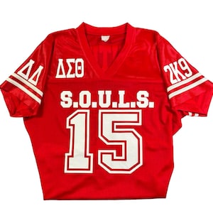 May include: Red football jersey with white lettering. The jersey has the letters "ΔΣΘ" on the left shoulder, "S.O.U.L.S." on the chest, and the number "15" on the front. The jersey also has the number "2K9" on the right shoulder.