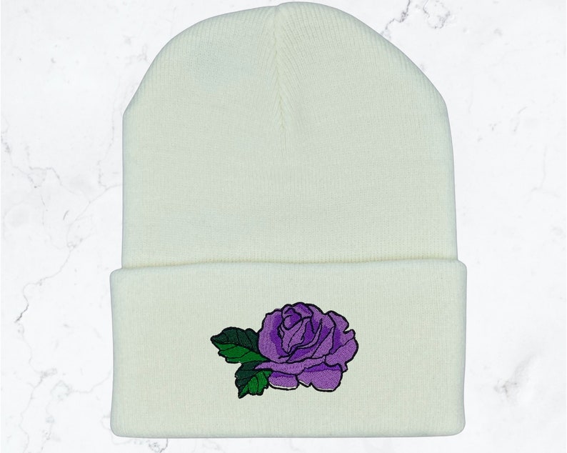 May include: White knitted beanie with a purple embroidered rose on the front.