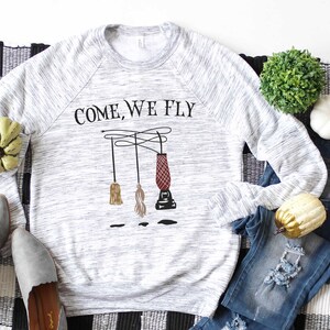 May include: A gray and white long-sleeved sweatshirt with the text "COME, WE FLY" printed on the front. The design features three broomsticks and a cauldron hanging from strings.