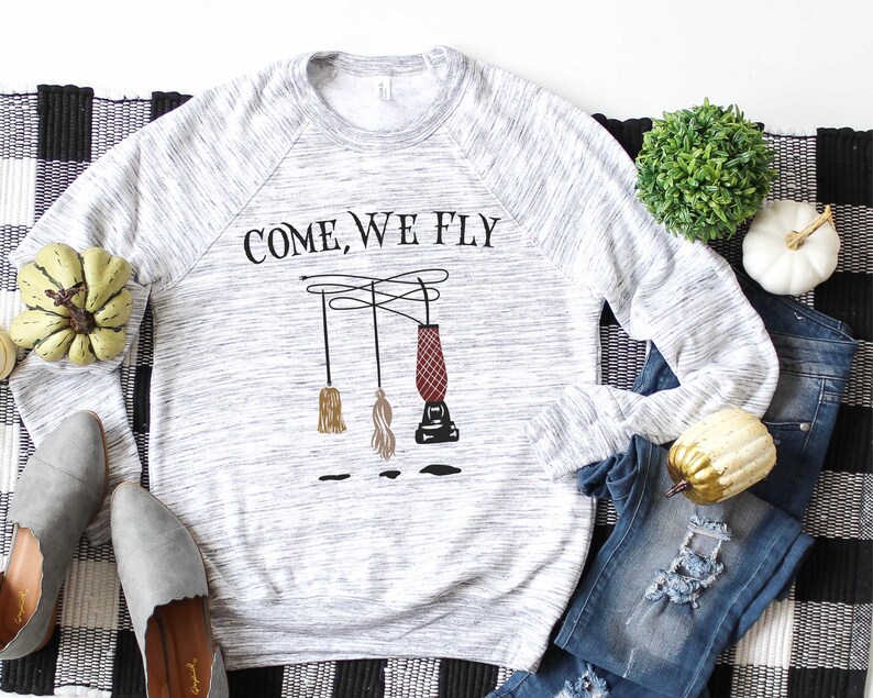 May include: A gray and white long-sleeved sweatshirt with the text "COME, WE FLY" printed on the front. The design features three broomsticks and a cauldron hanging from strings.
