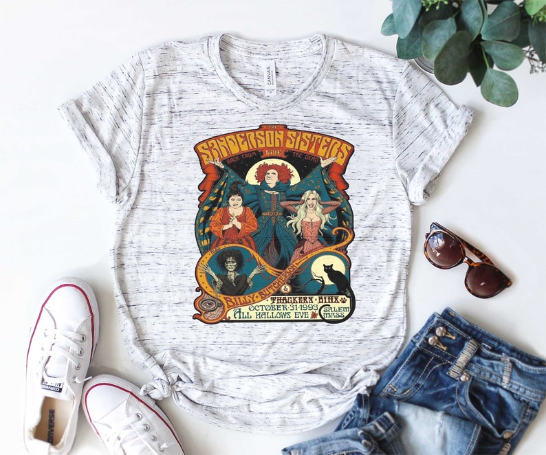 Pu&ograve; includere: Una maglietta bianca con una grafica in stile vintage delle sorelle Sanderson del film Hocus Pocus. La grafica presenta le tre streghe, un gatto nero e il testo "The Sanderson Sisters Back From The Dead Billy Butcherson Thackery Binx October 31, 1993 Salem All Hallows Eve".