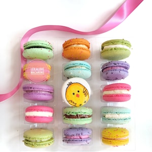 May include: A box of 12 pastel-colored macarons with a chick design on one. The macarons are arranged in a clear plastic box with a pink ribbon on top. The box has a label that says "LeilaLove Macarons".