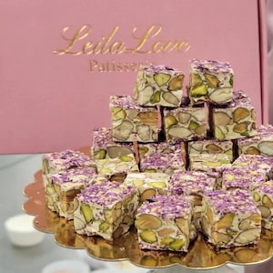 May include: A pyramid of square nougat pieces topped with purple flower petals and filled with whole pistachios and almonds. The treats are arranged on a gold scalloped tray in front of a pink box labeled Leila Love Patisserie.