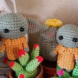 Op de afbeelding: Twee gehaakte groene en oranje Yoda-achtige figuren met grote ogen, zittend in groene en oranje cactuspotten.