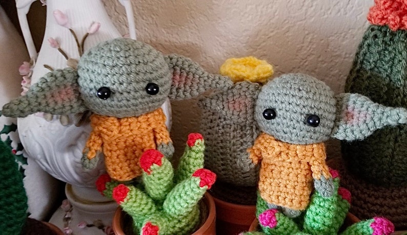 Op de afbeelding: Twee gehaakte groene en oranje Yoda-achtige figuren met grote ogen, zittend in groene en oranje cactuspotten.