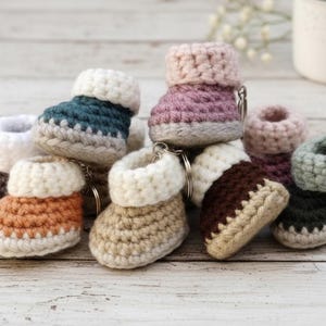 May include: A collection of handmade crochet baby bootie keychains in various colors, including teal, orange, pink, and brown. Each bootie features a white cuff and a silver keychain ring. The booties are arranged on a light wooden surface.