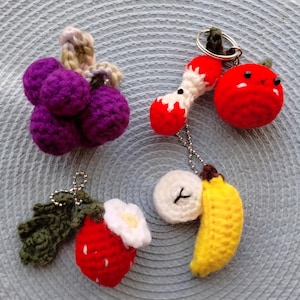 May include: A collection of handmade crochet keychains featuring various fruits and vegetables. Includes a bunch of purple grapes, a red tomato, a red strawberry with a white flower, a yellow banana, and a white onion.