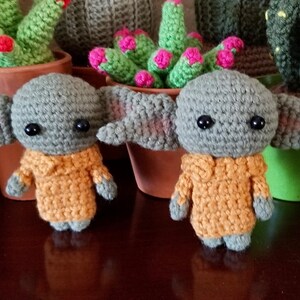 Op de afbeelding: Twee gehaakte groene en gele pluche speelgoedjes die lijken op een populair fictief personage. De speelgoedjes staan op een houten oppervlak.