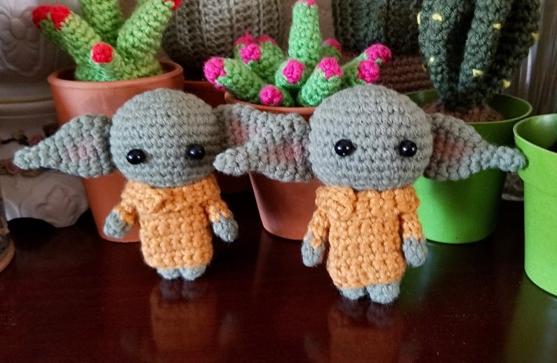 Op de afbeelding: Twee gehaakte groene en gele pluche speelgoedjes die lijken op een populair fictief personage. De speelgoedjes staan op een houten oppervlak.