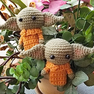 Op de afbeelding: Twee gehaakte groene en oranje pluche speelgoedjes die lijken op een populair fictief personage. De speelgoedjes zitten in een pot met groene bladeren.