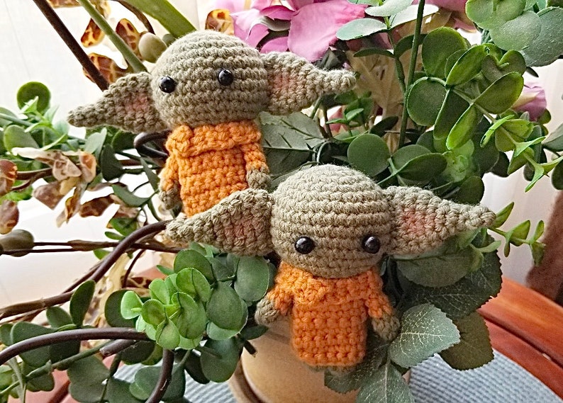 Op de afbeelding: Twee gehaakte groene en oranje pluche speelgoedjes die lijken op een populair fictief personage. De speelgoedjes zitten in een pot met groene bladeren.