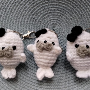 May include: Three white crocheted seal keychains with black eyes and noses. Each seal has a black bow on top of its head. The keychains are hanging from a silver metal ring.
