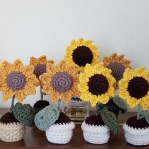 May include: A collection of crocheted sunflower decorations in various shades of yellow, brown, and green. Each sunflower has a crocheted pot with a brown, beige, or white base.