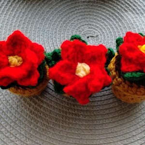 May include: Three handmade crochet poinsettia flowers. Each flower has vibrant red petals, a yellow center, and green leaves. The flowers are set in small, tan-colored pots, perfect for holiday decor.