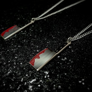 May include: Two silver necklaces with a silver cleaver pendant. The cleaver has a red drip design on the blade.