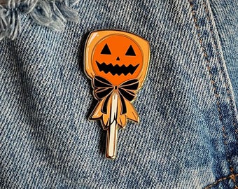 Op de afbeelding: Een Halloween-thema emaille pin in de vorm van een pompoen lolly. De pin heeft een oranje pompoengezicht met een zwarte strik en een witte stok. De pin is geplaatst tegen een denim achtergrond.