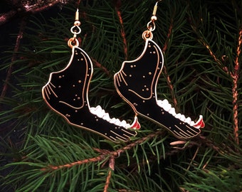 May include: A pair of gold earrings shaped like a black animal jaw with white teeth. The earrings are hanging from a branch.