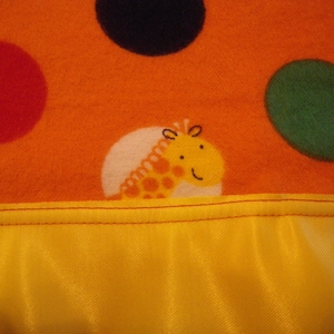 May include: An orange baby blanket with large, colorful polka dots and a yellow satin trim. A cartoon giraffe is featured on the blanket. The blanket is made of soft material and has red stitching.